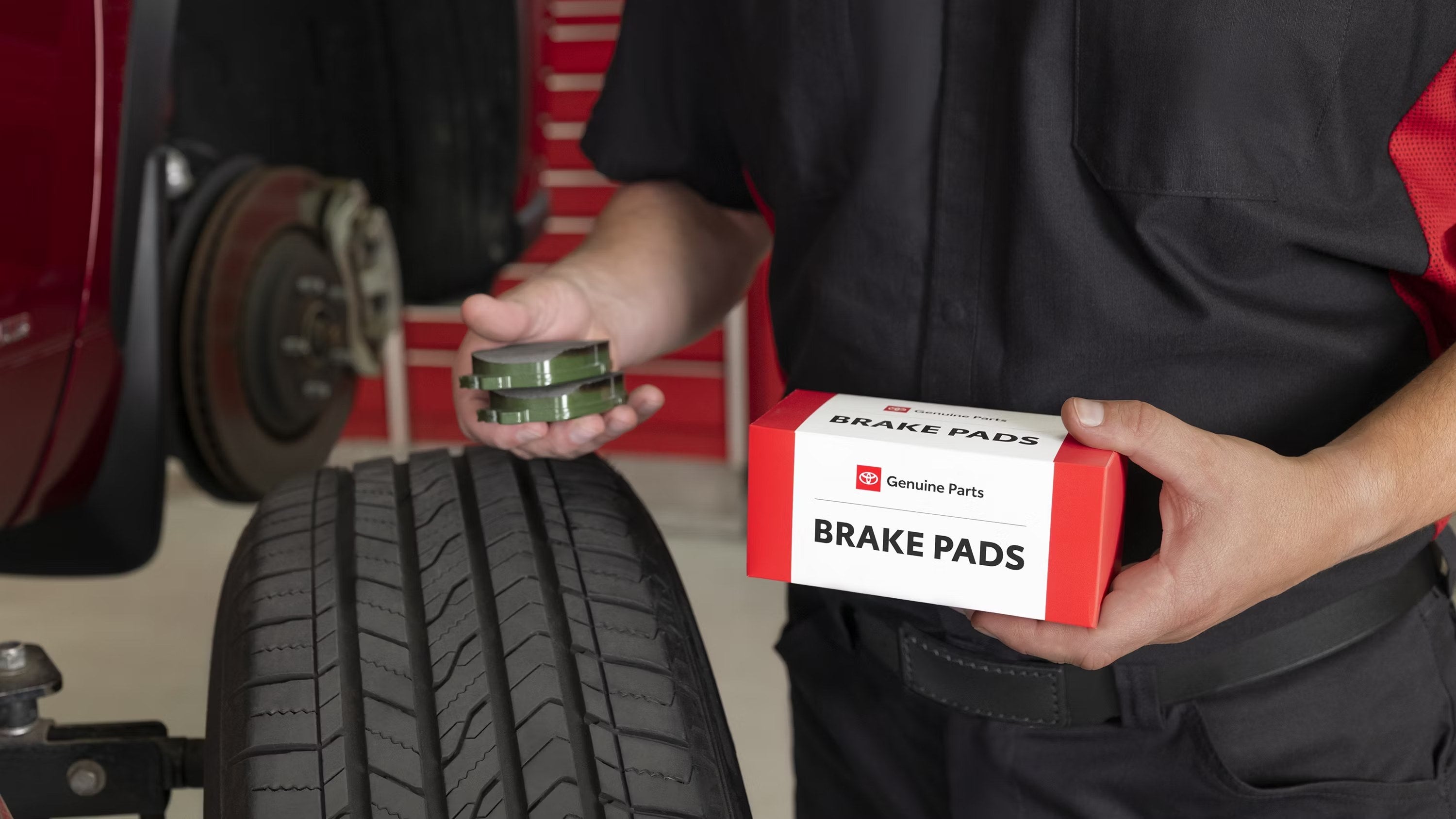 Technician holding a Toyota Genuine Parts brake pad box and a set of brake pads next to a wheel